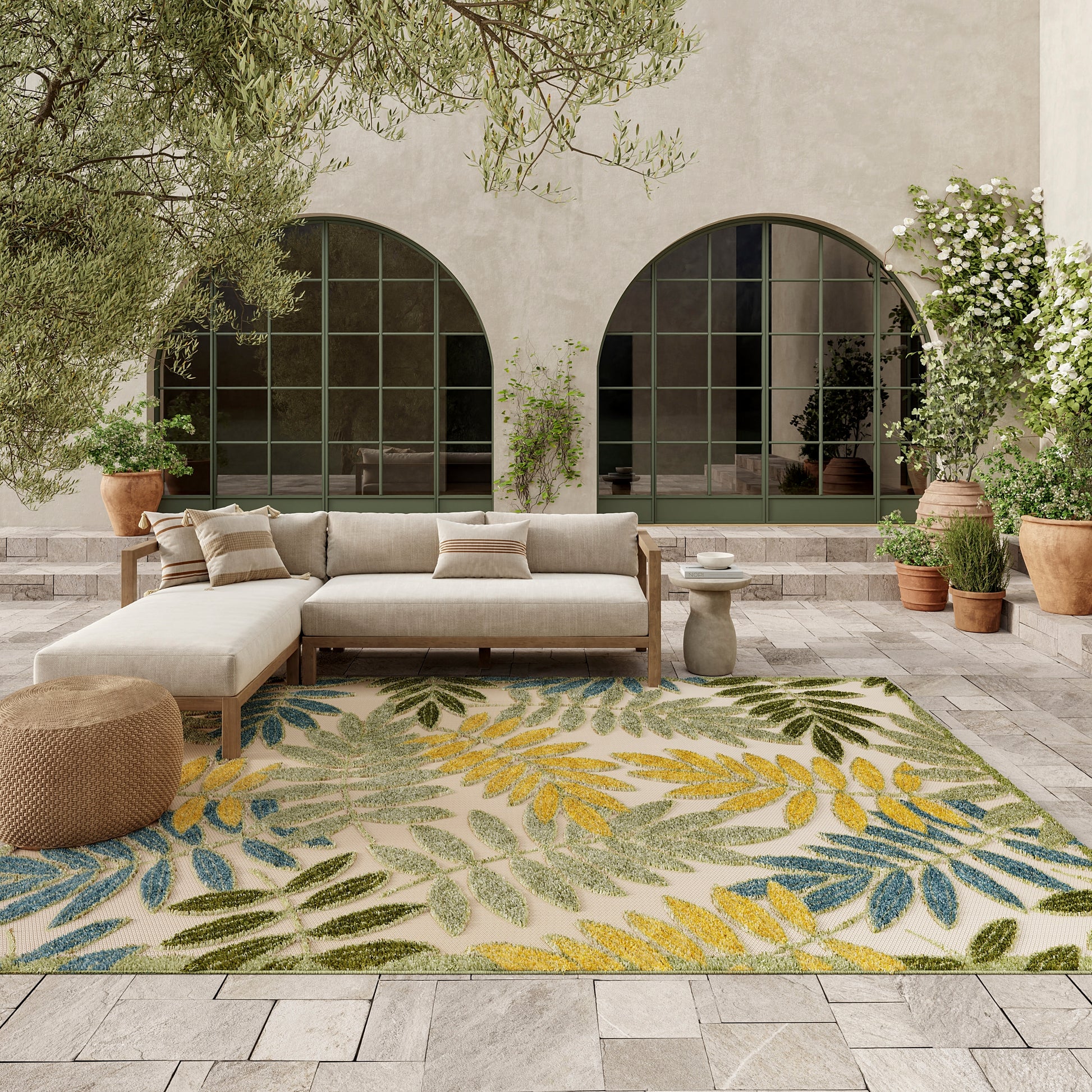 Outdoor courtyard seating with beige sectional sofa, tropical leaf rug in blue/green/yellow, potted plants & arched windows.