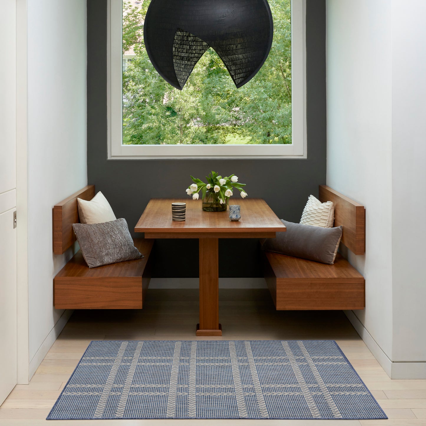 Wooden corner dining nook with built-in benches and square table, black pendant lamp, blue rug, cushions, flowers in vase.