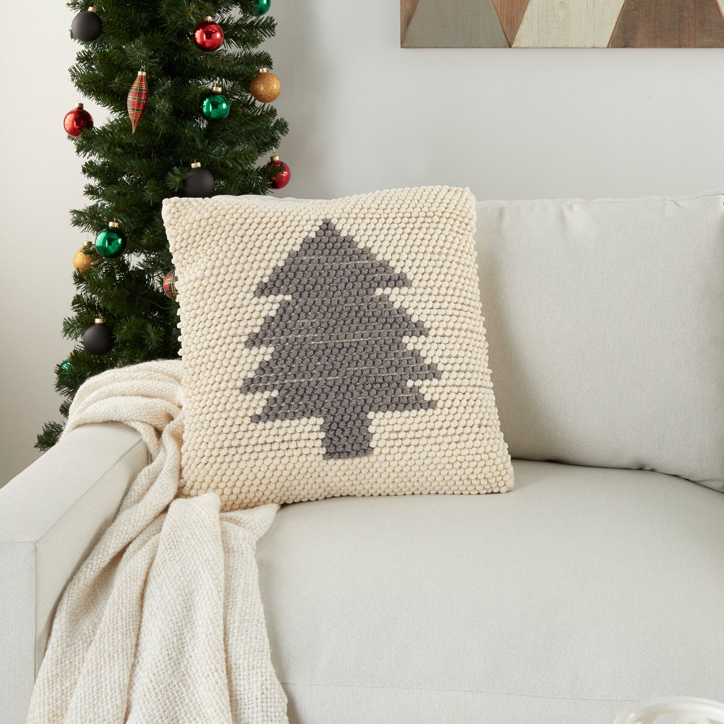 Cream chunky-knit pillow with a gray Christmas tree motif on a beige sofa beside a decorated Christmas tree.