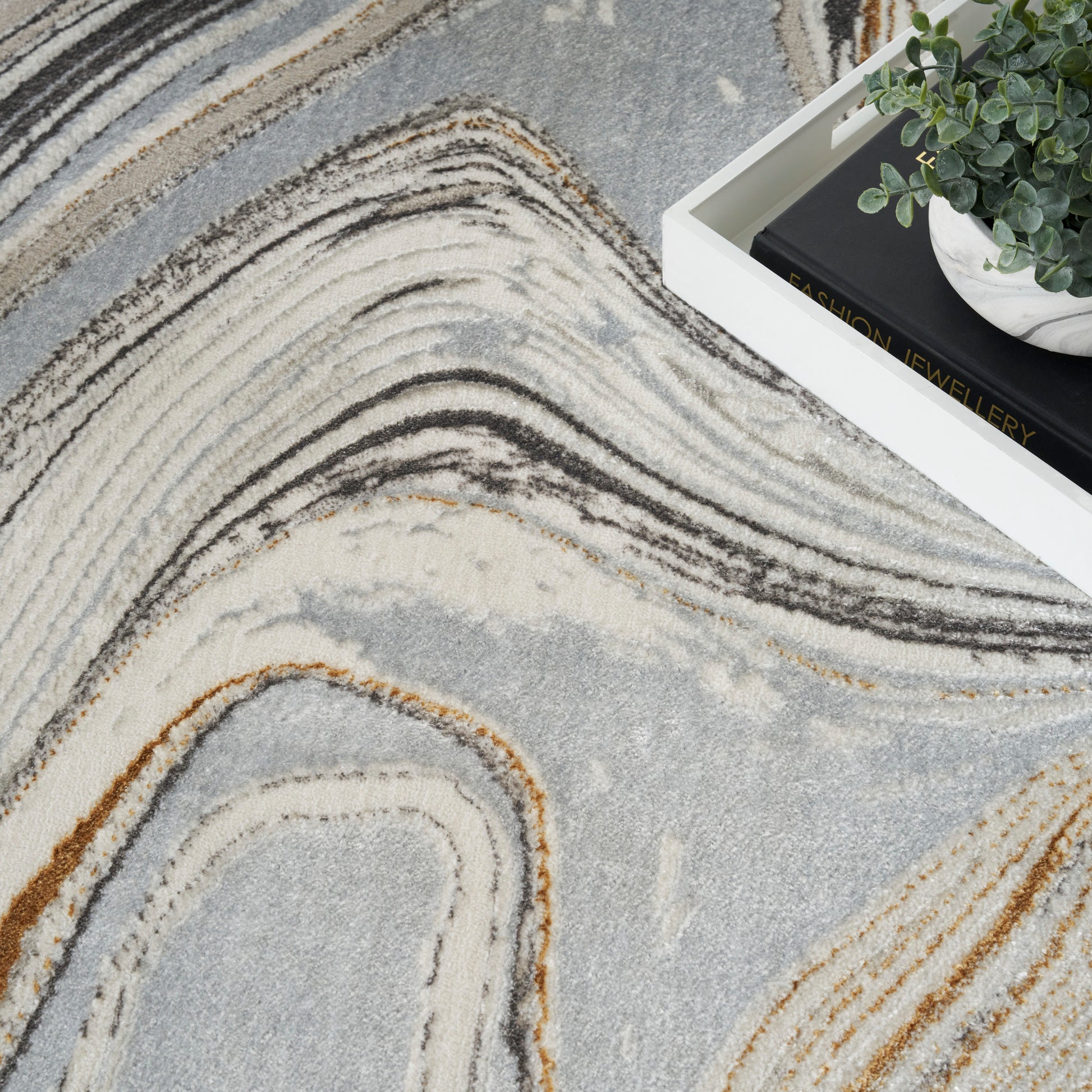 Grey abstract rug with cream, beige & brown swirls; white tray with plant & a fashion jewellery book.
