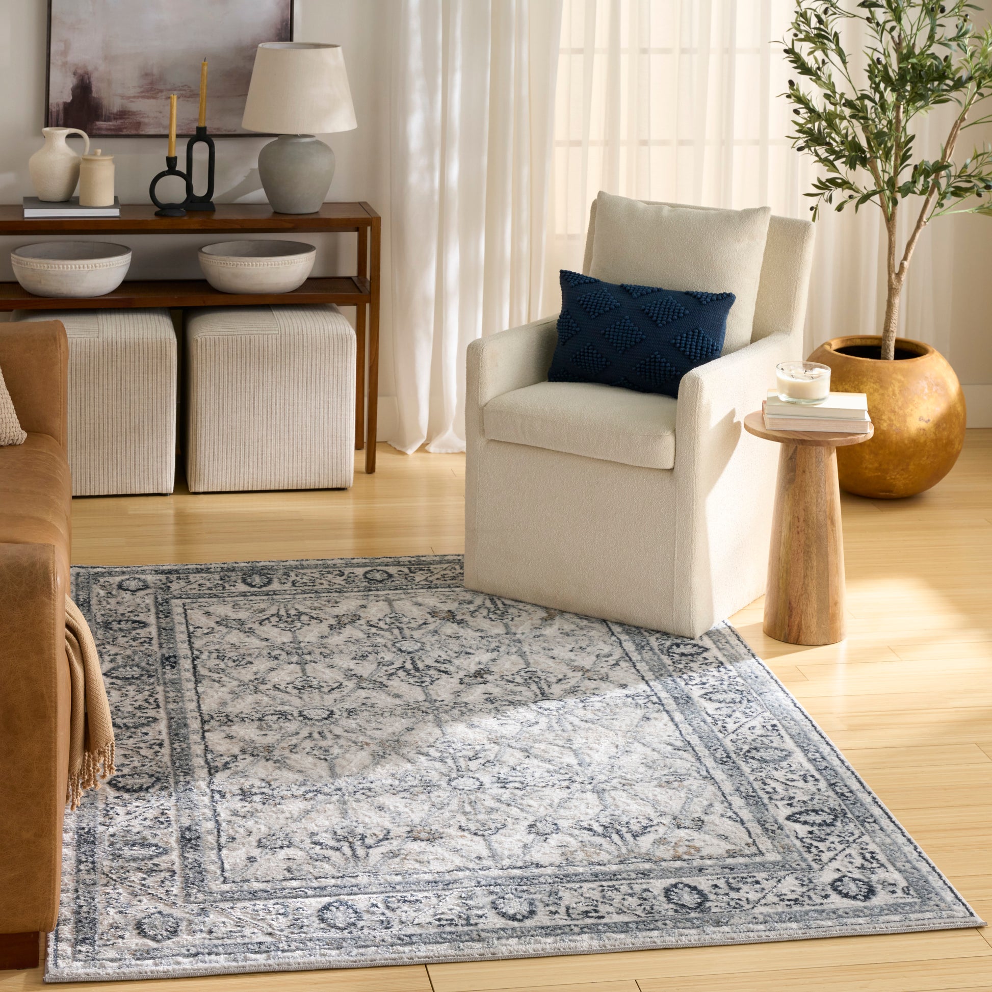 Beige fabric armchair with navy textured pillow, wooden side table, potted tree, and blue-gray patterned area rug.