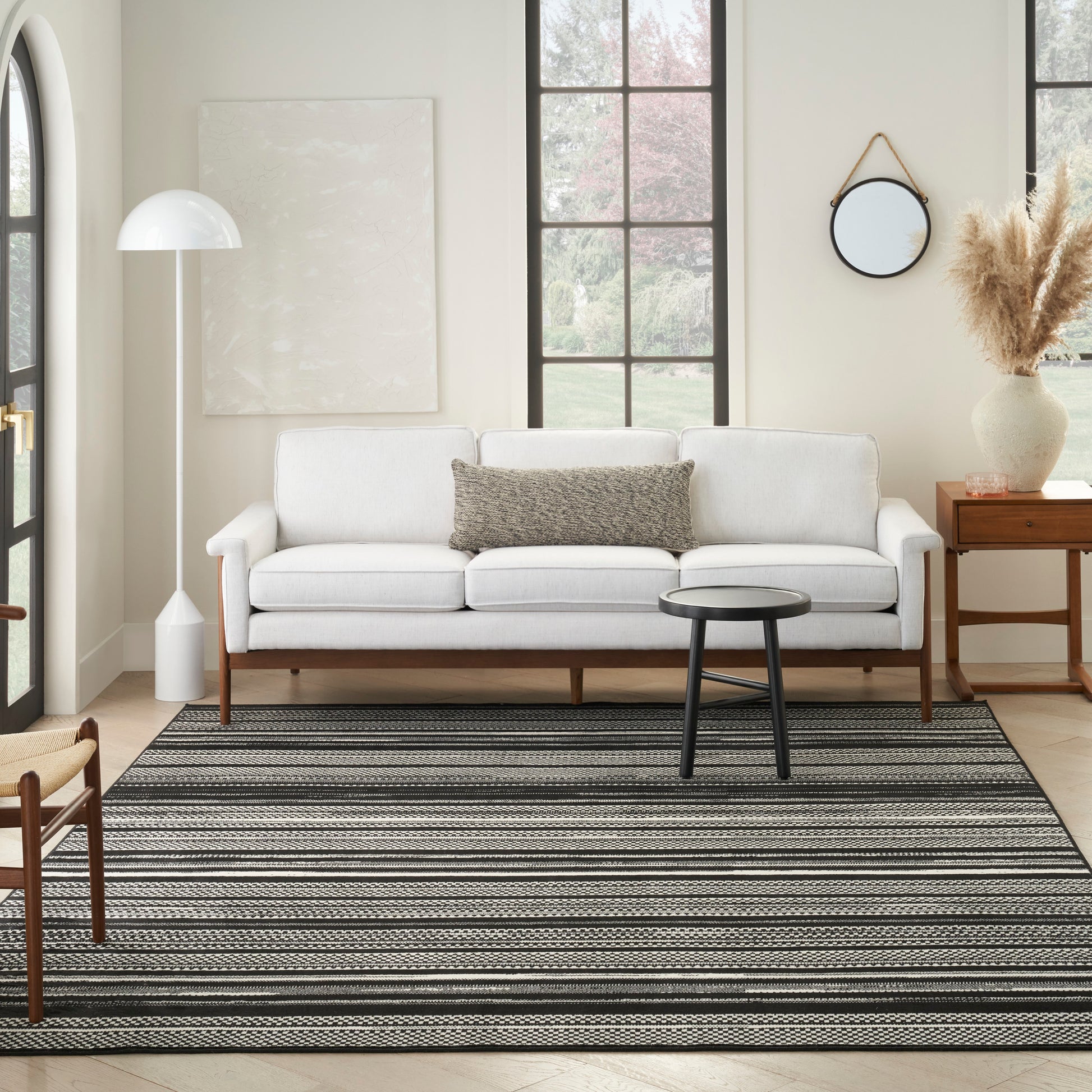 Bright living room with white sofa, gray cushion, black round coffee table, striped grey-black rug, floor lamp, pampas vase.