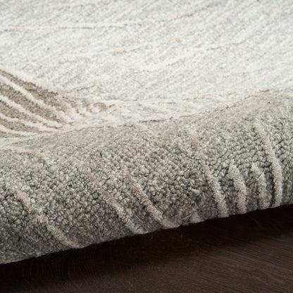 Close-up of a neutral gray-beige rug with plush cut-pile texture and rolled edge on dark wood.