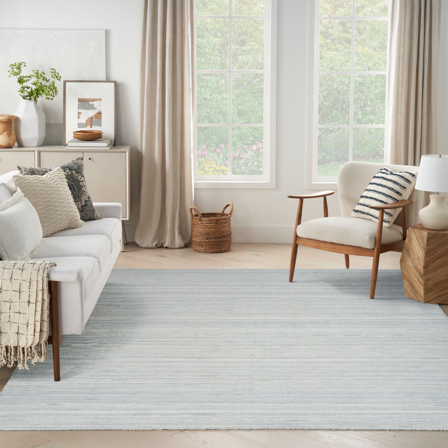 Bright living room with pale sofa, cream armchair, light blue rug, woven basket, large windows with beige curtains.