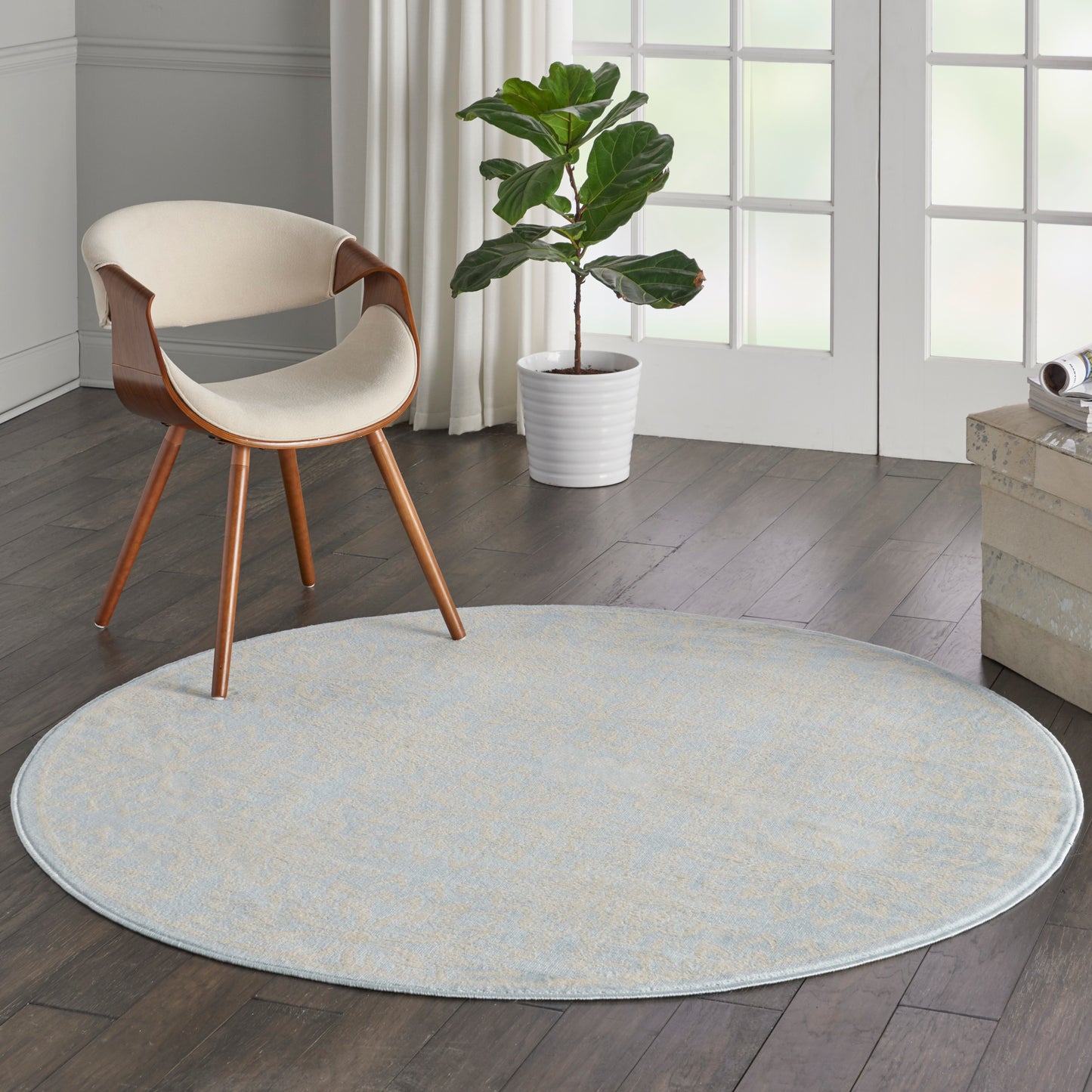 Round pale blue rug on a wood floor with a cream curved-seat chair and wooden legs beside a tall plant in a white pot.
