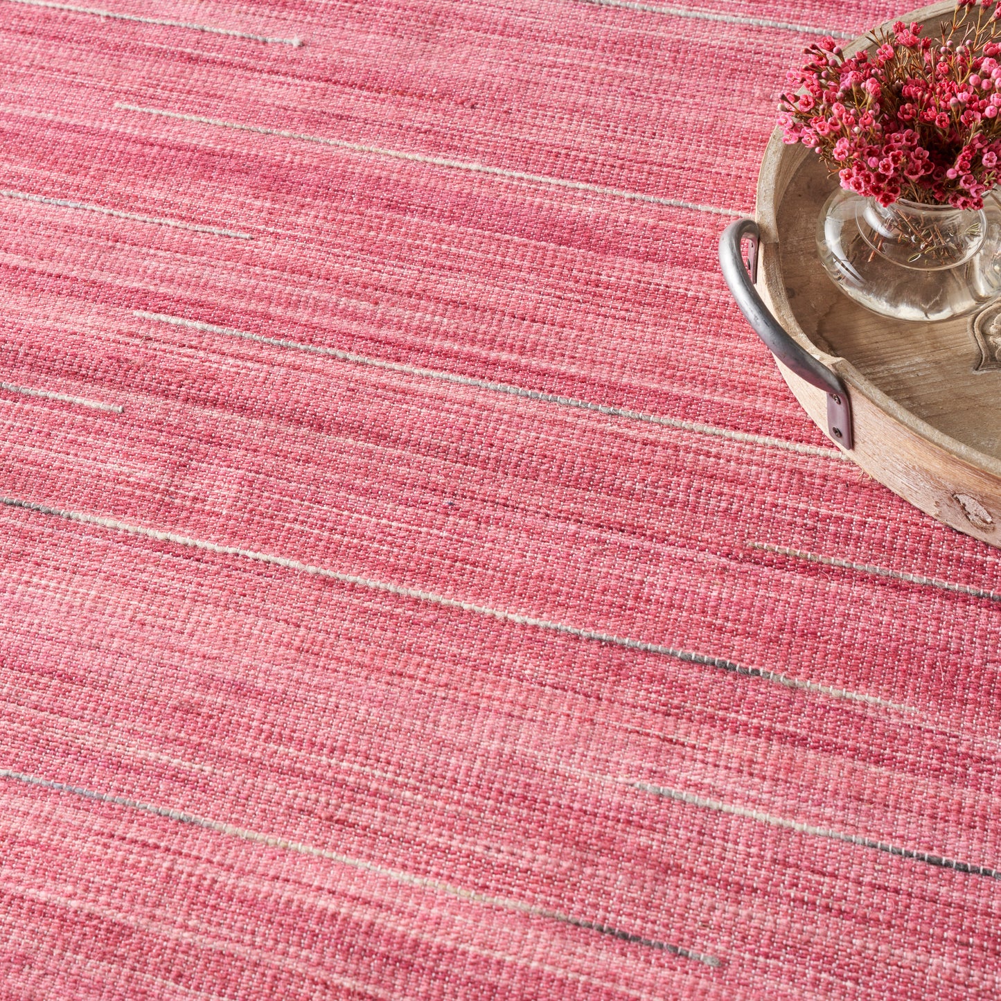Pink striped woven rug with a round wooden tray holding a glass vase of pink flowers.