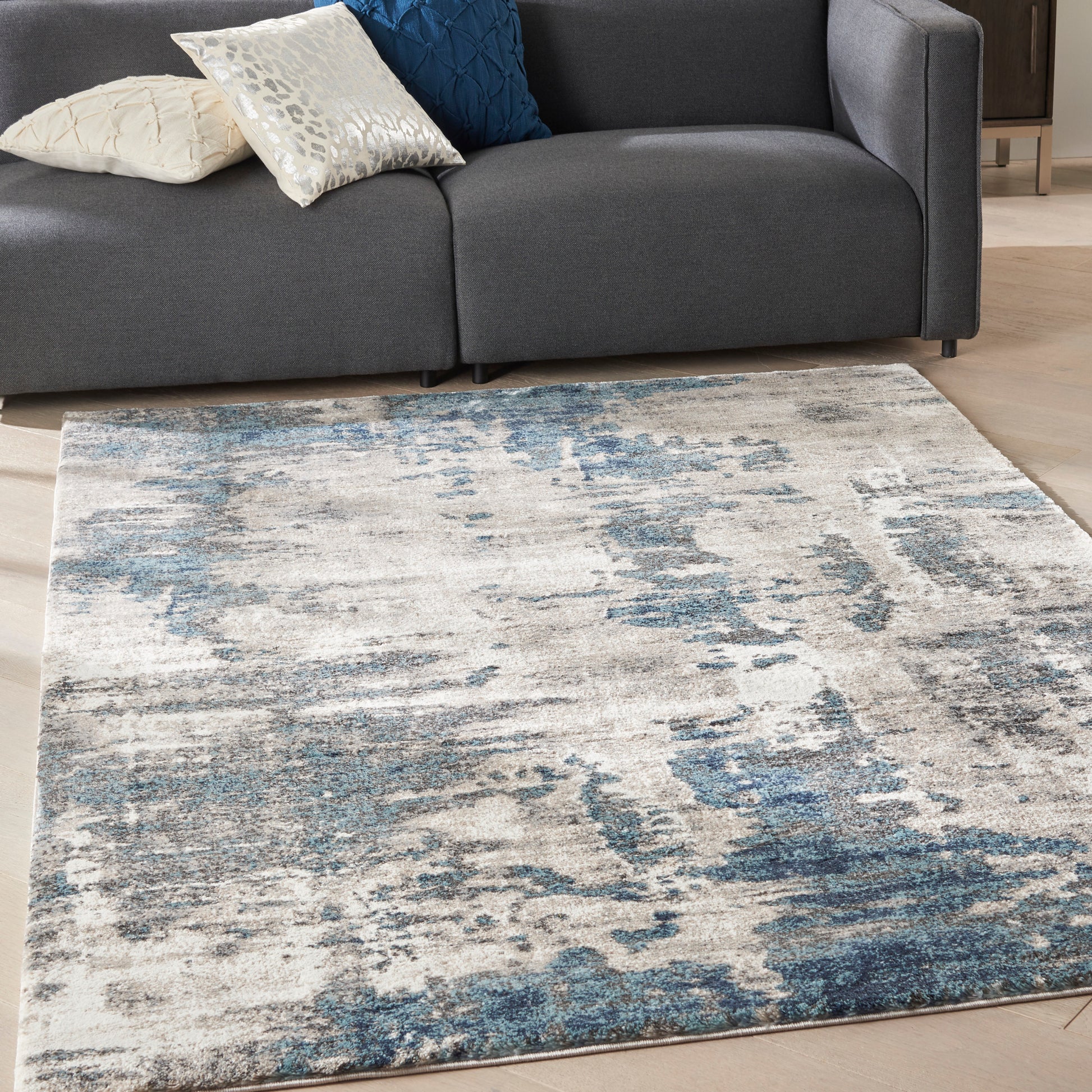 Distressed abstract blue-ivory area rug 5' x 7' on a light wood floor beside a dark gray sectional with blue and cream pillows.