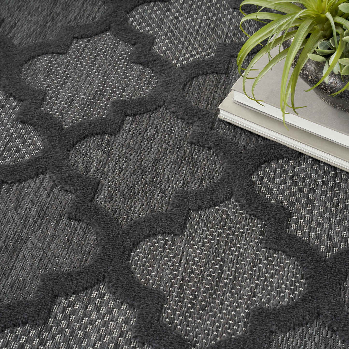 Charcoal geometric trellis rug with interlocking shapes, dark gray weave, beside a plant and stacked magazines.