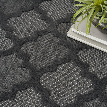 Charcoal geometric trellis rug with interlocking shapes, dark gray weave, beside a plant and stacked magazines.
