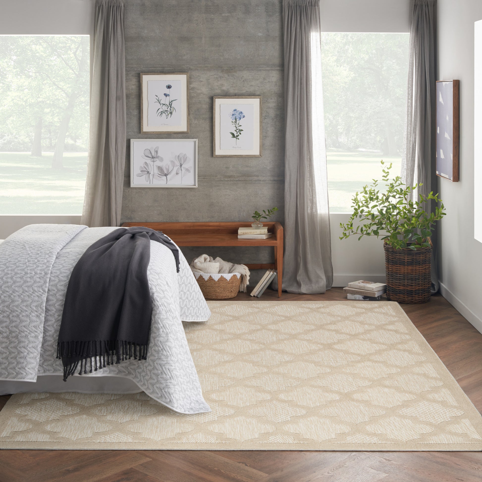 Neutral bedroom with beige geometric rug, white quilt, gray throw, concrete wall, botanical art, wooden bench, plants.
