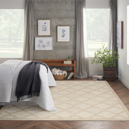 Neutral bedroom with beige geometric rug, white quilt, gray throw, concrete wall, botanical art, wooden bench, plants.