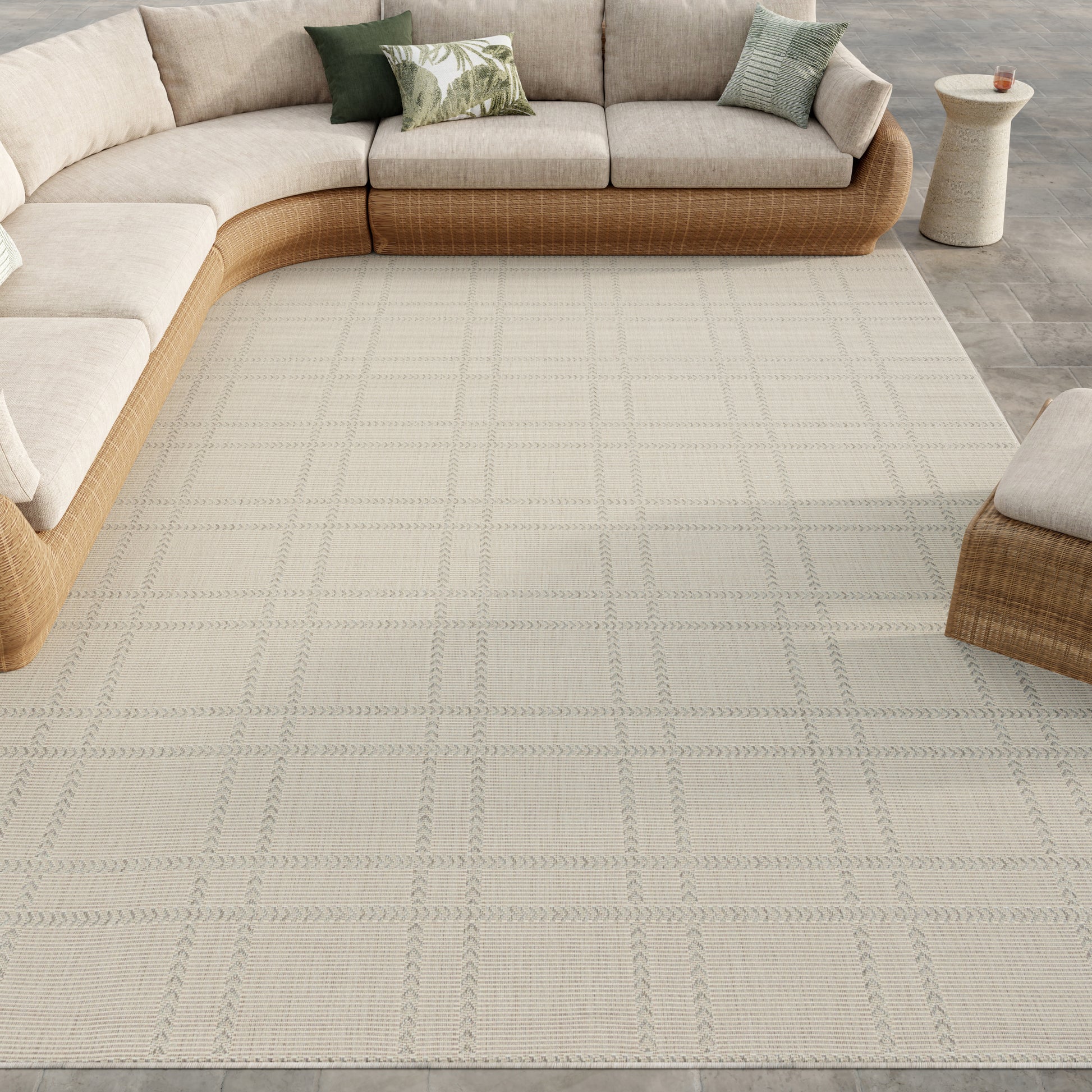 Beige sectional with woven wicker base, light grid-pattern rug, tropical green pillows, and a small beige patio side table.