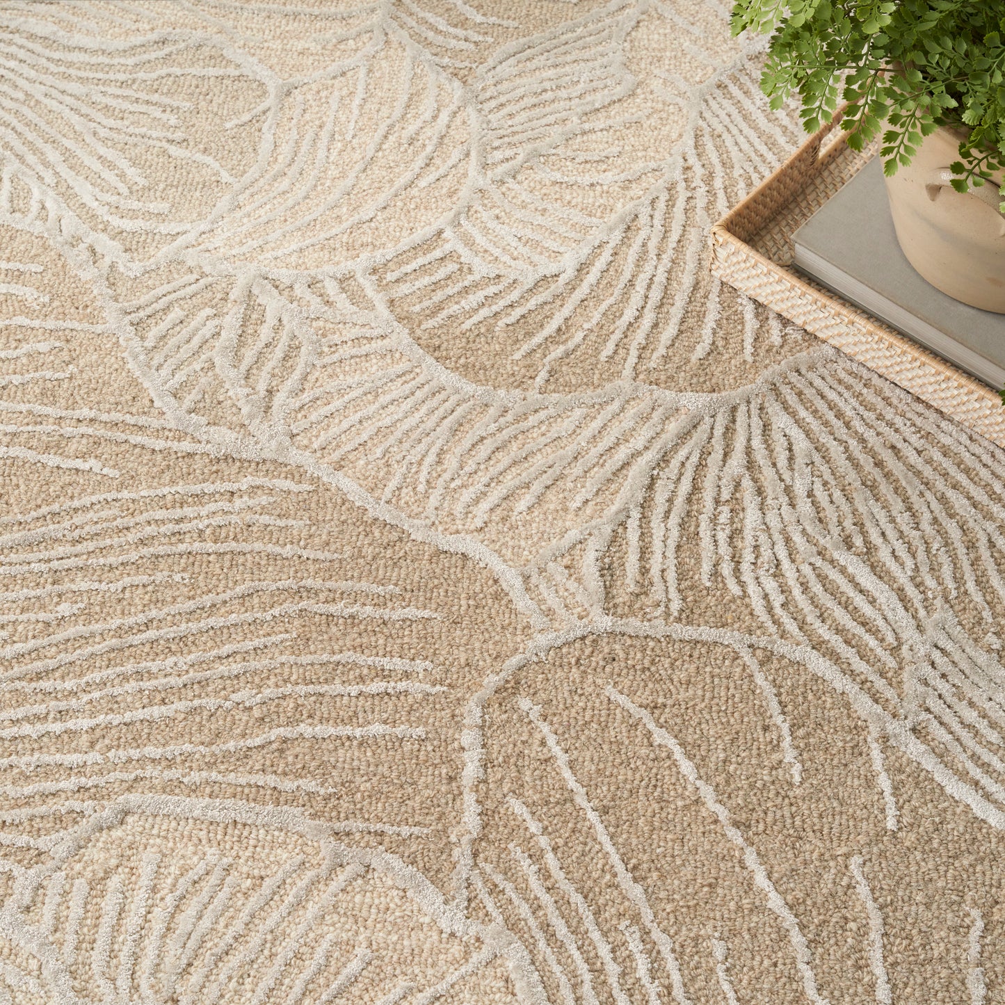 Beige area rug with cream palm-leaf pattern; soft textured shag, shown with a wooden tray and plant in the corner.