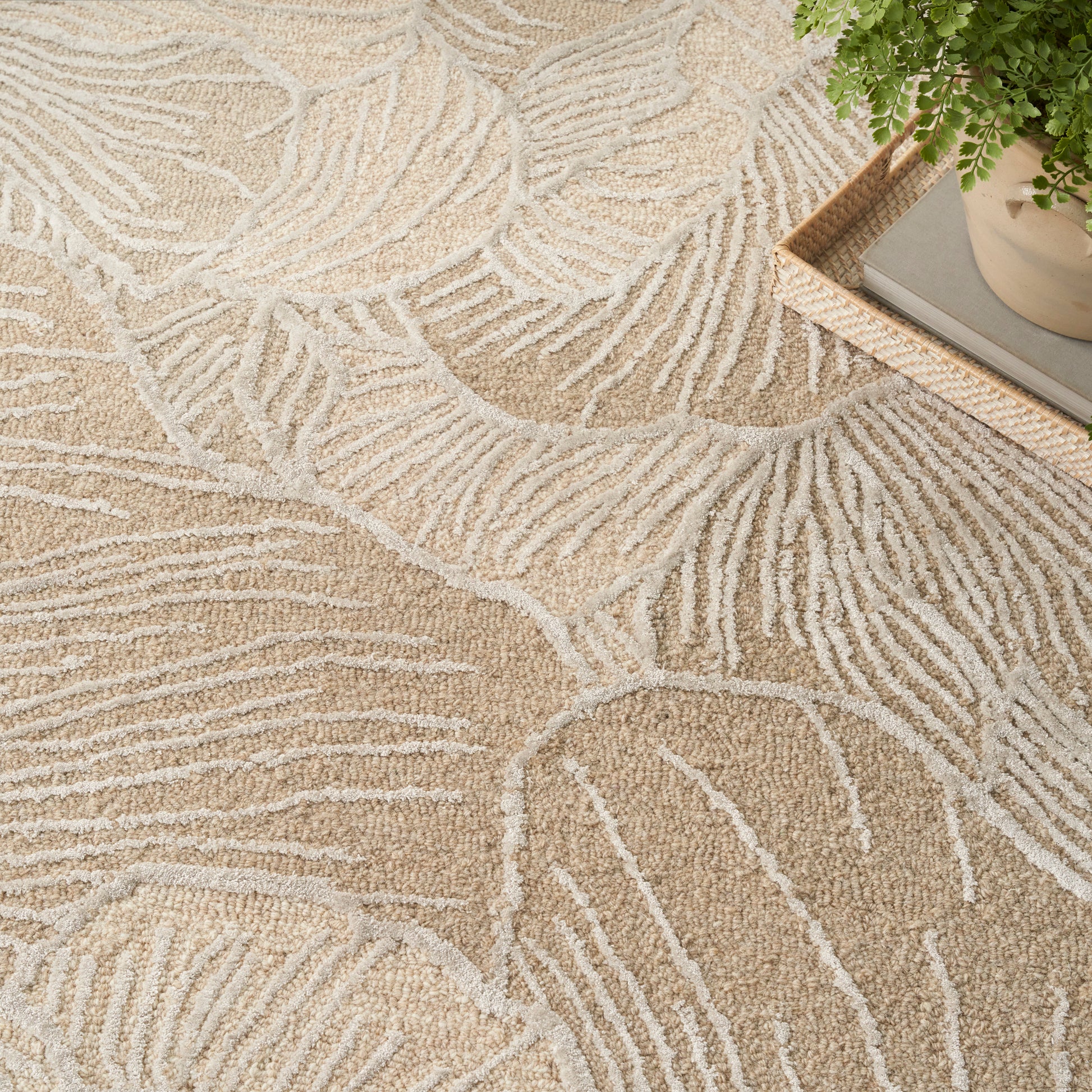 Beige area rug with cream palm-leaf pattern; soft textured shag, shown with a wooden tray and plant in the corner.
