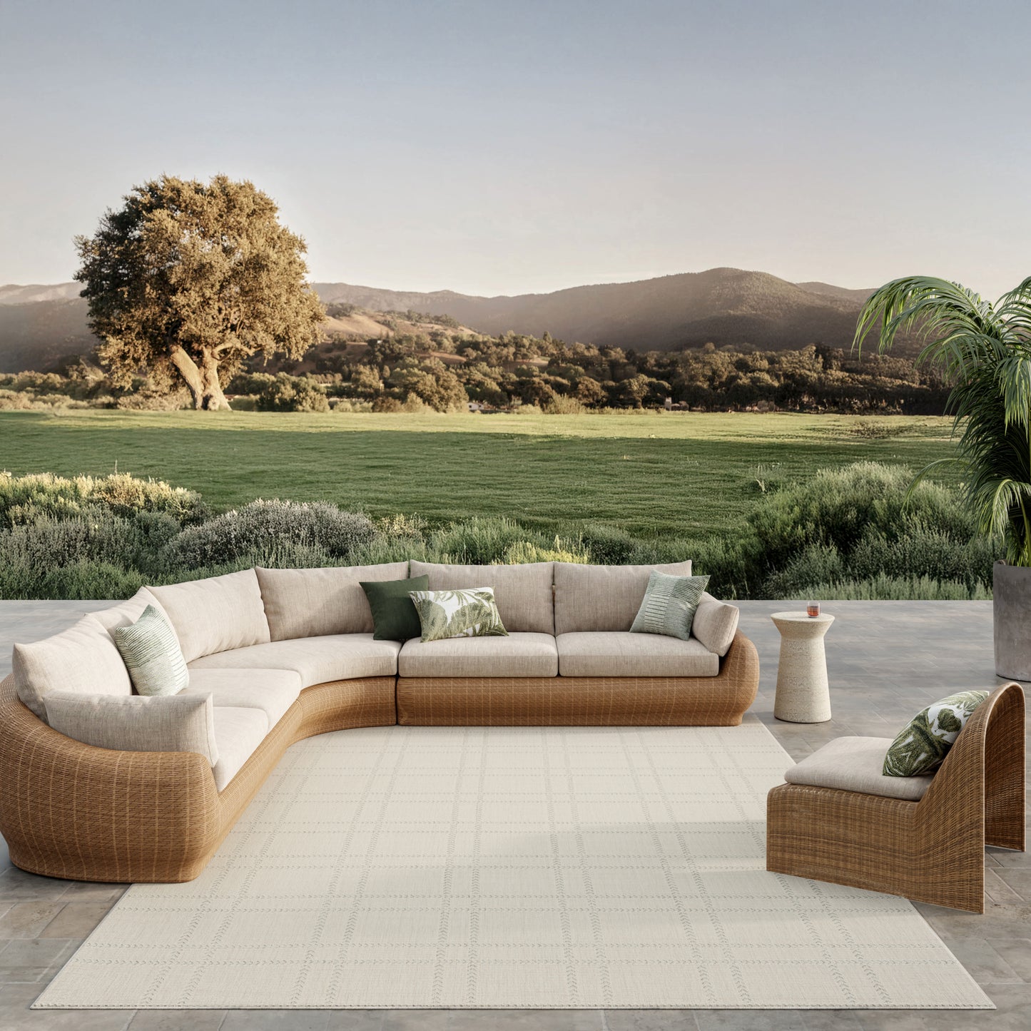 Outdoor rattan sectional sofa with beige cushions and green throw pillows on a light rug, scenic lawn and hills backdrop.