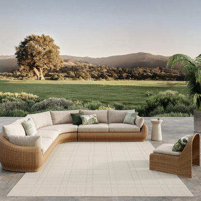 Outdoor rattan sectional sofa with beige cushions and green throw pillows on a light rug, scenic lawn and hills backdrop.