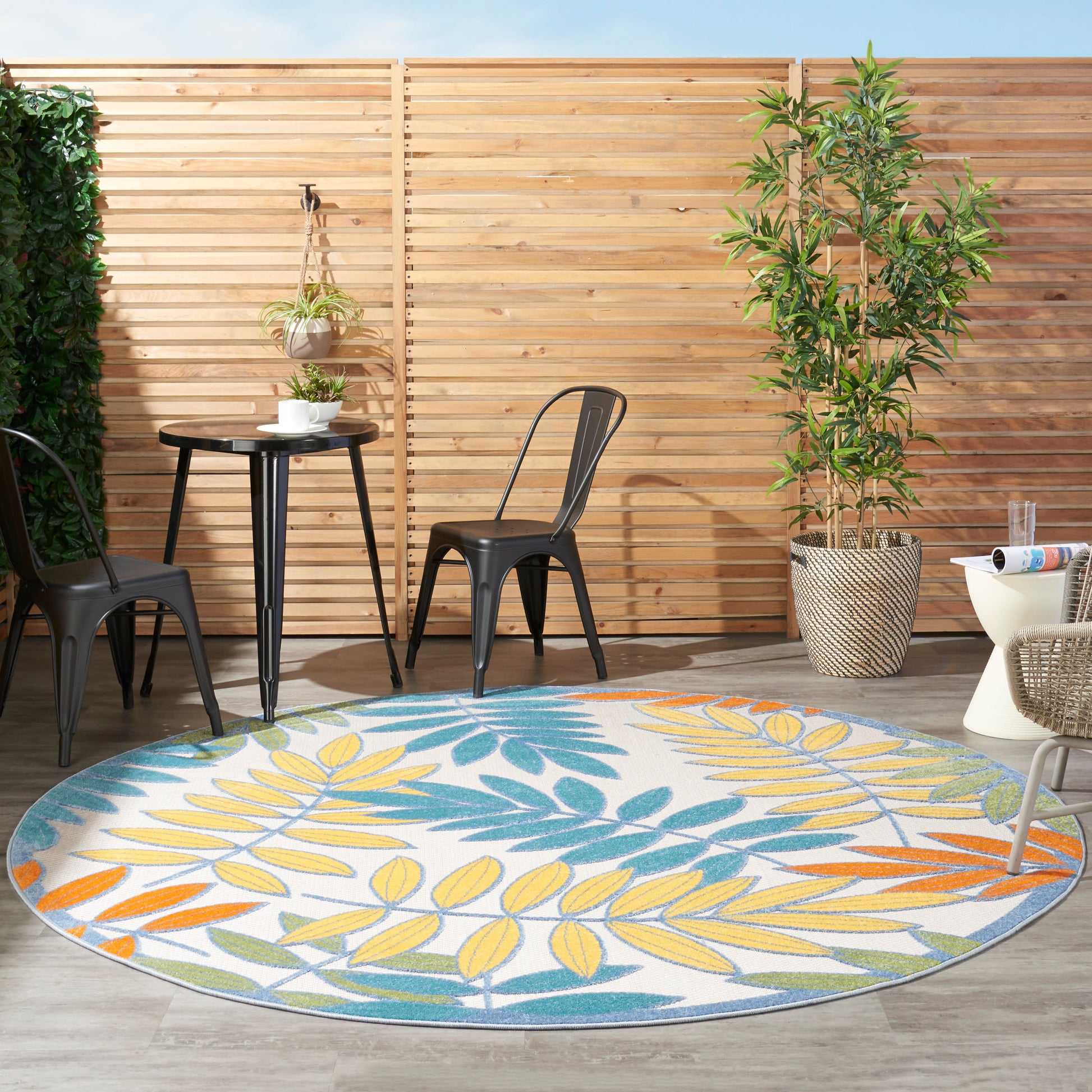 Round outdoor rug with yellow, teal, blue & orange leaf pattern; black metal chairs & table on a fence with plants.