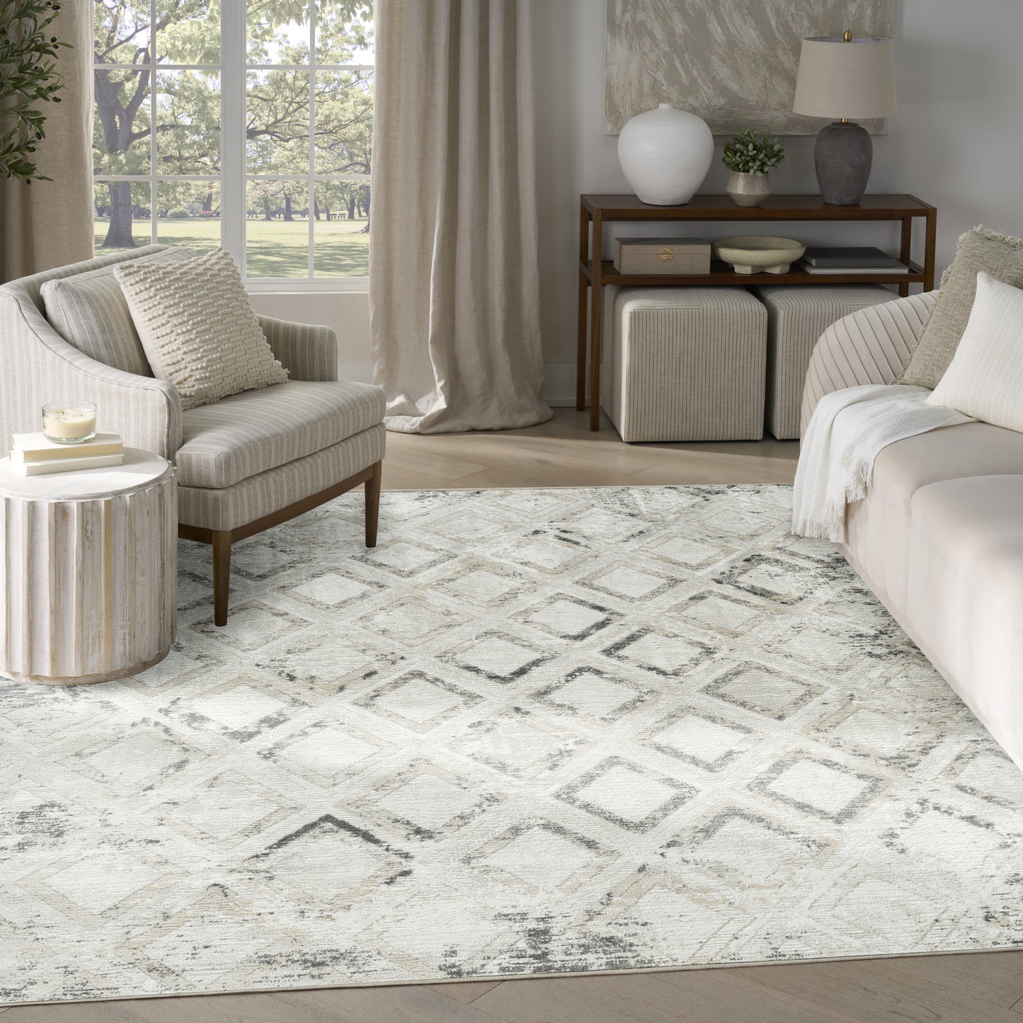 Neutral living room with cream sofa, beige armchair, diamond rug, light wood furniture and large window with greenery.