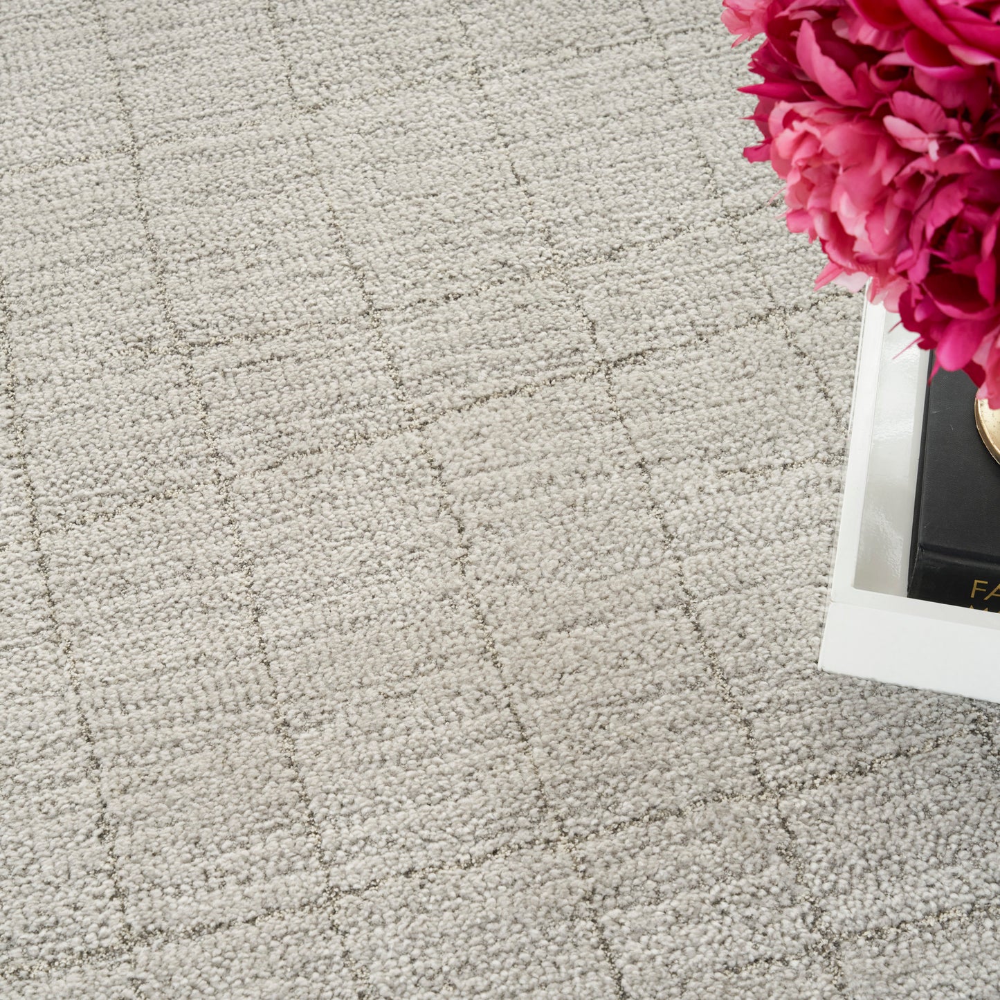 Beige textured rug with a subtle grid pattern and a white tray holding bright pink peonies in the top-right corner.