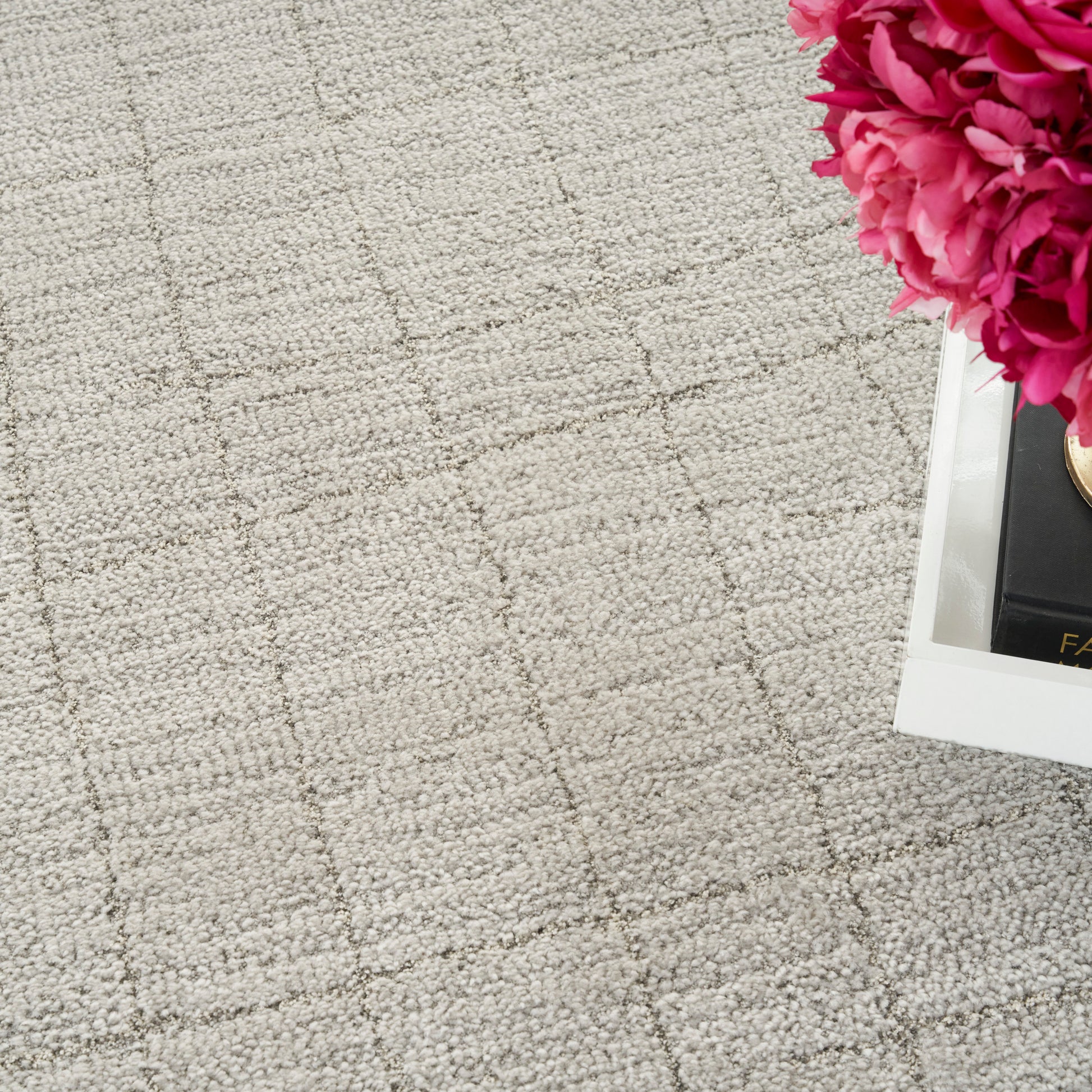 Beige textured rug with a subtle grid pattern and a white tray holding bright pink peonies in the top-right corner.