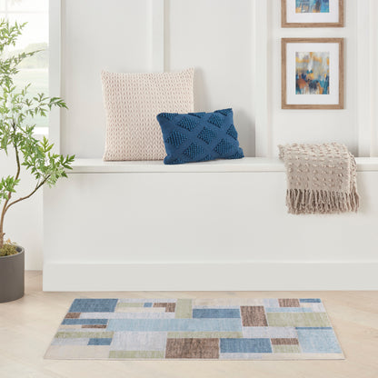 Cozy seating nook with beige knit pillow, blue diamond-pattern pillow, fringed throw, multicolor patchwork rug, plant.