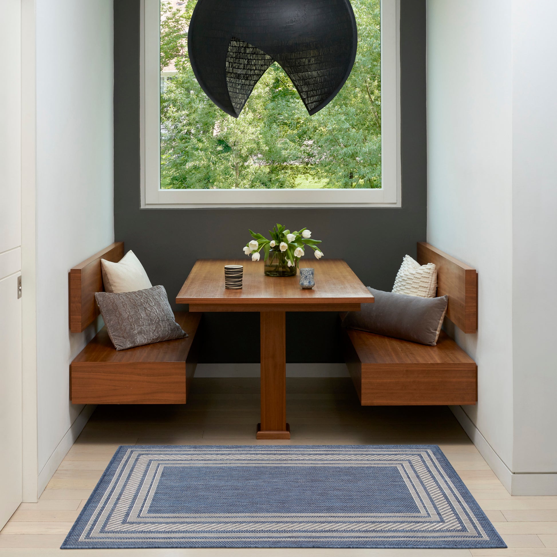Wooden corner dining nook with built-in benches, square table, gray walls, black curved pendant, blue striped rug, vase of flowers.