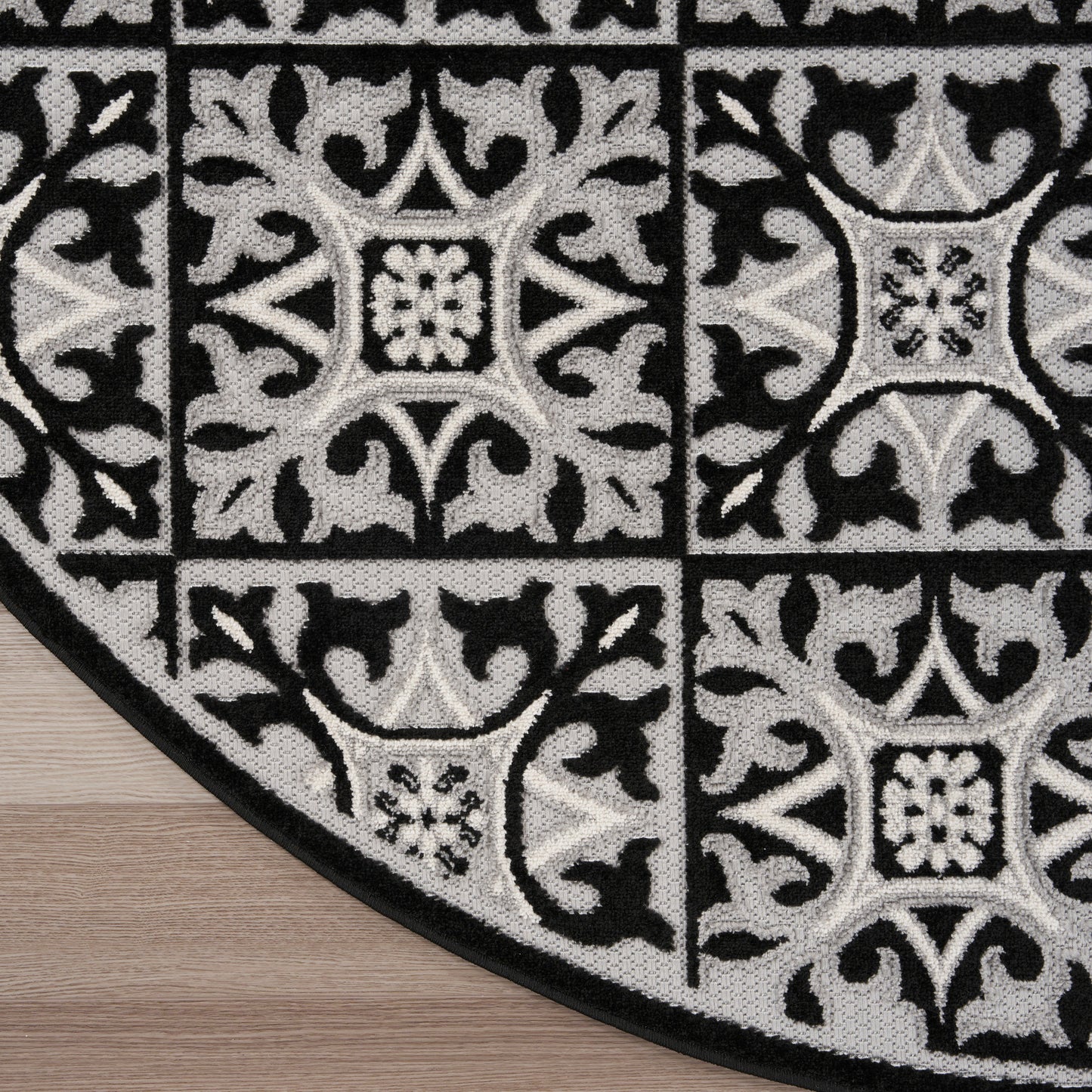Round black & gray rug with tile-like square medallions and ornate floral scrollwork on wood floor.