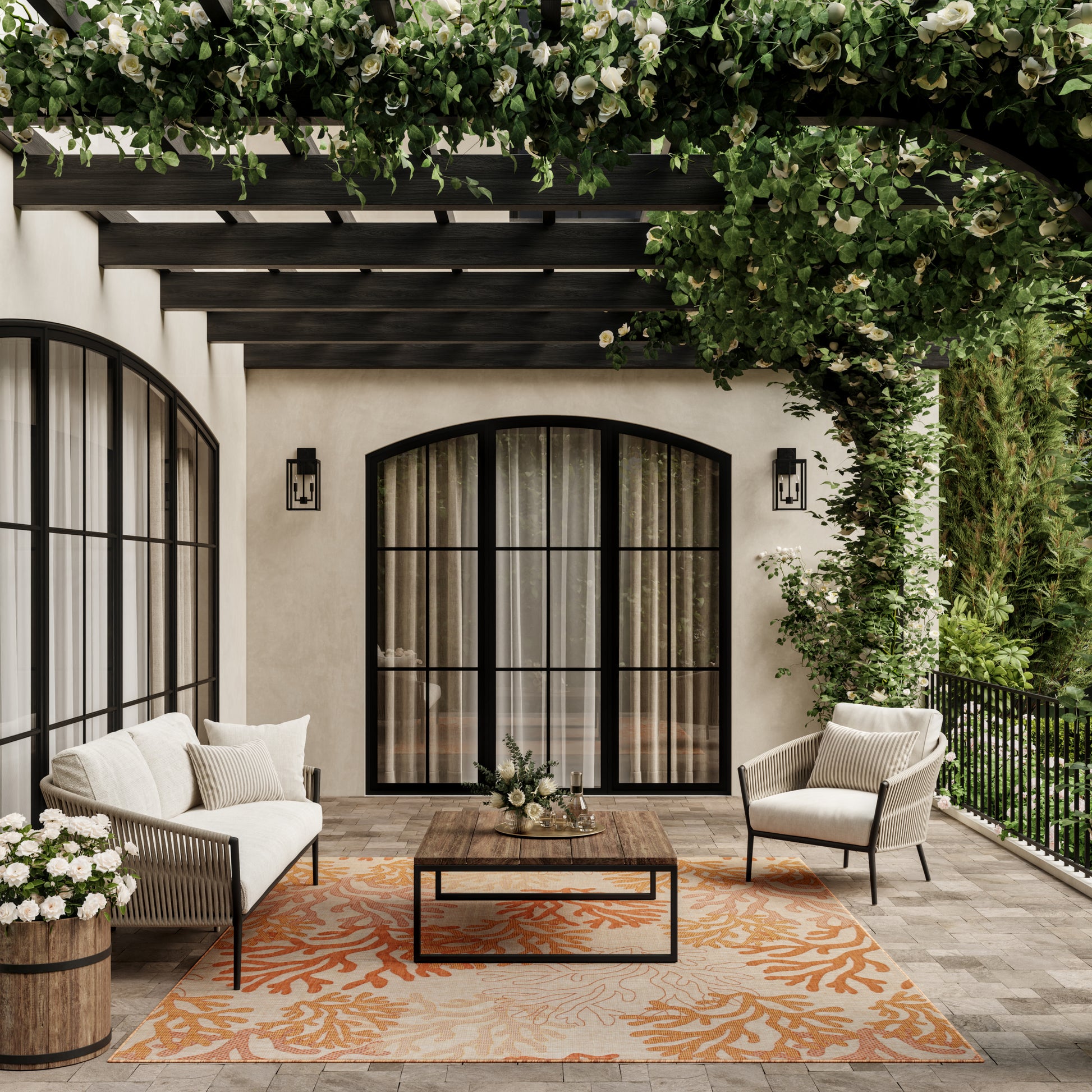 Cozy outdoor patio with beige stucco, black arched doors, cream seating, wood coffee table, orange coral rug, and lush greenery.