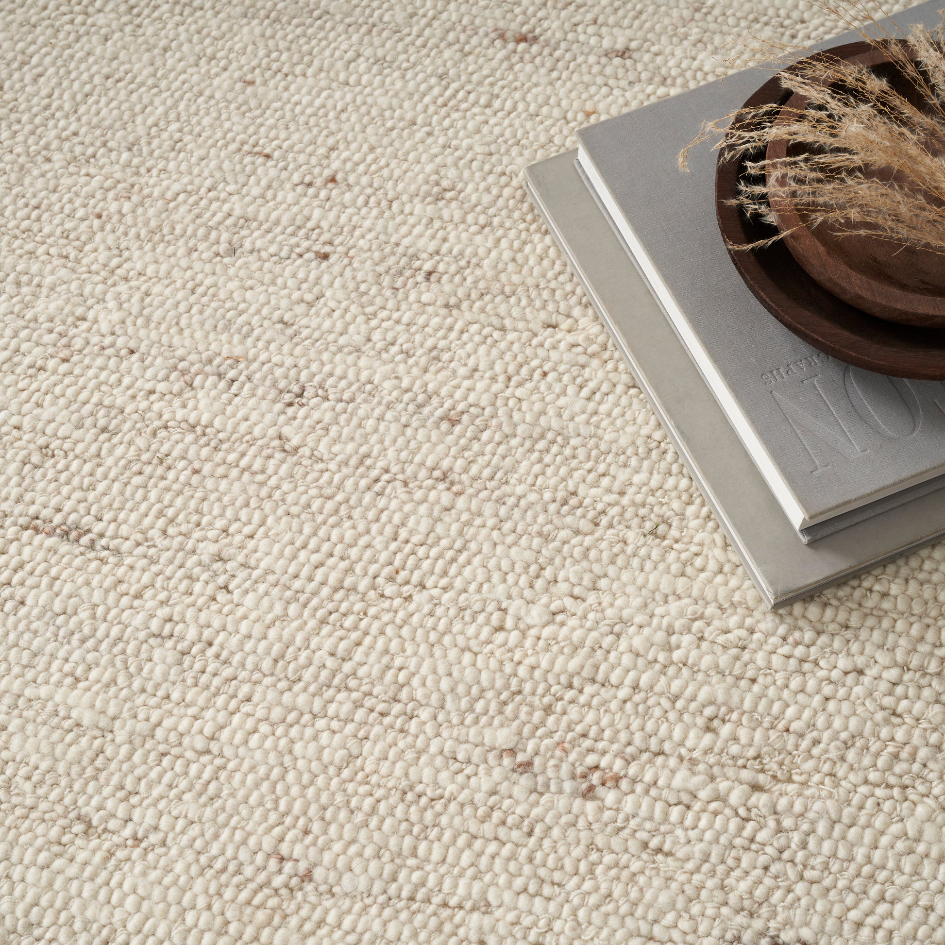 Beige textured woven rug with a stack of gray magazines and a round wooden dish holding dried grasses.