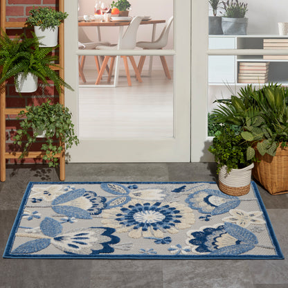 Outdoor rectangular rug with blue floral pattern on beige background and blue border at the entrance beside potted plants.