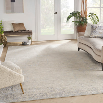 Bright living room with large neutral rug, cream sofa, beige ottoman, wooden bench, potted plants, and glass doors.