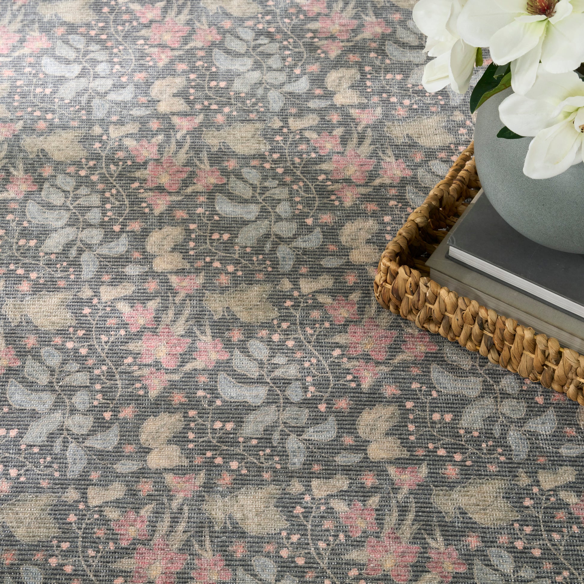 Floral gray-pink woven rug with vine pattern; wicker tray, gray vase, white flowers, and books.