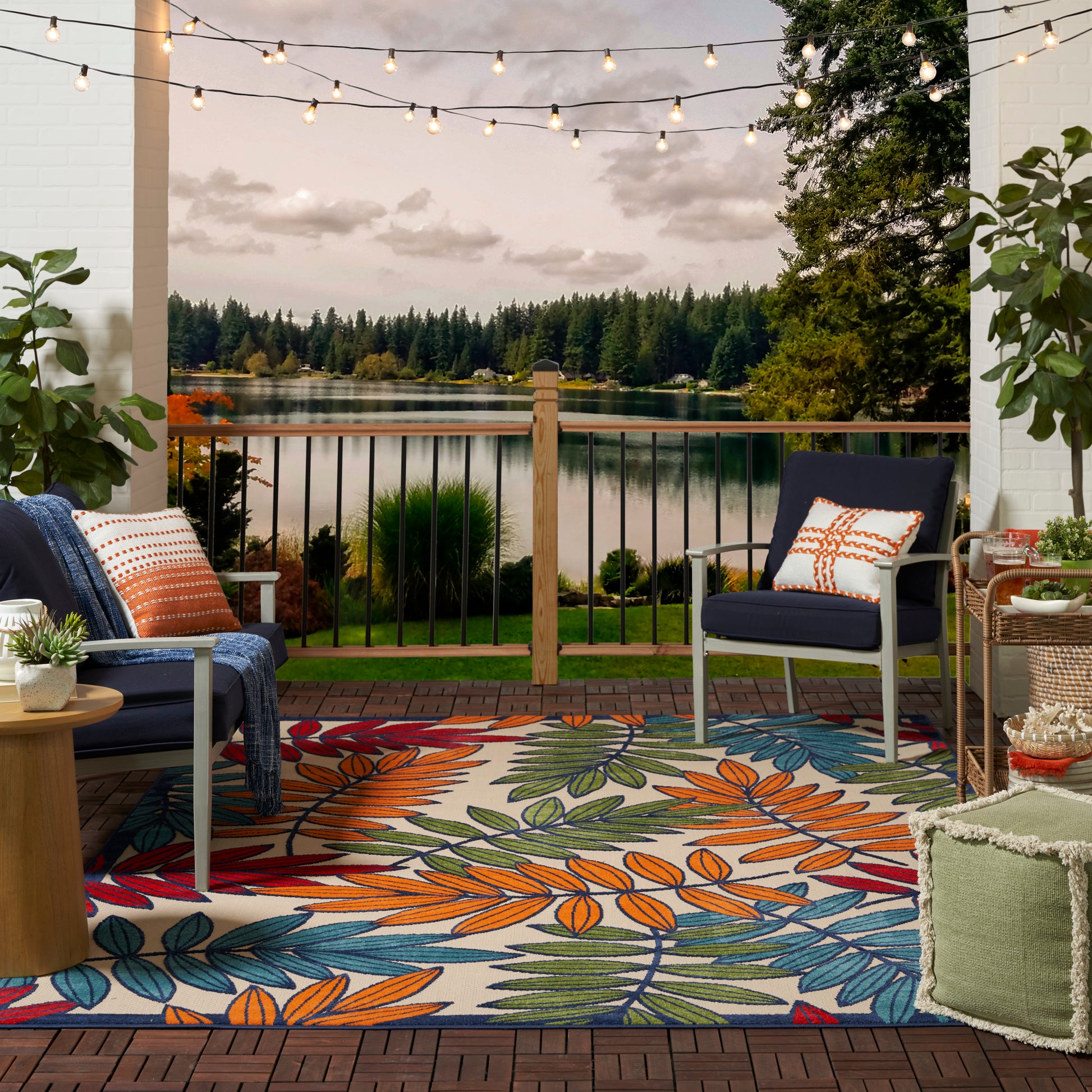 Cozy outdoor porch by a lake at dusk with colorful leaf-pattern rug, navy chairs, orange/white pillows, and string lights.
