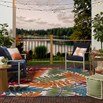 Cozy outdoor porch by a lake at dusk with colorful leaf-pattern rug, navy chairs, orange/white pillows, and string lights.