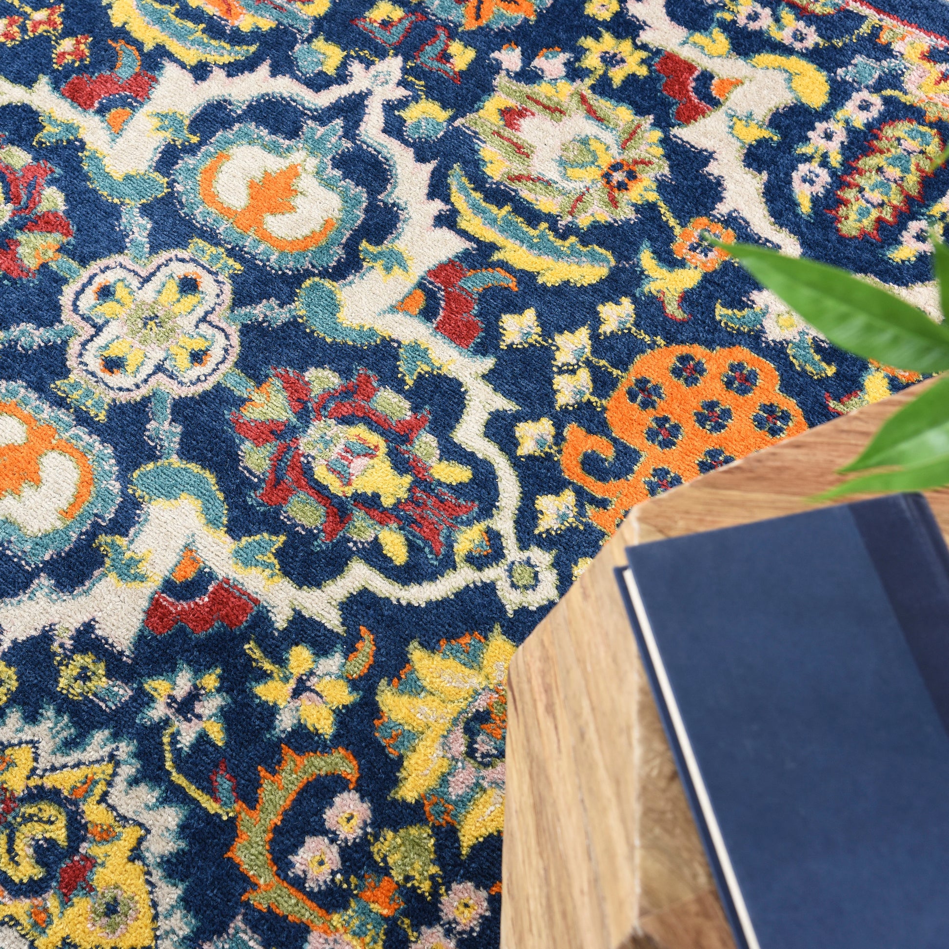Colorful Bohemian-inspired rug with a navy base and floral patterns; wooden table edge, blue book, and green plant nearby.