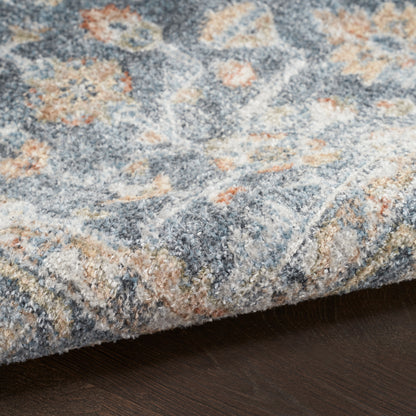 Blue-gray floral area rug with peach and ivory accents; edge folded over on dark wood floor.