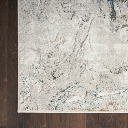 Rectangular cream-gray abstract area rug with blue veining and white fringe on dark wood floor.