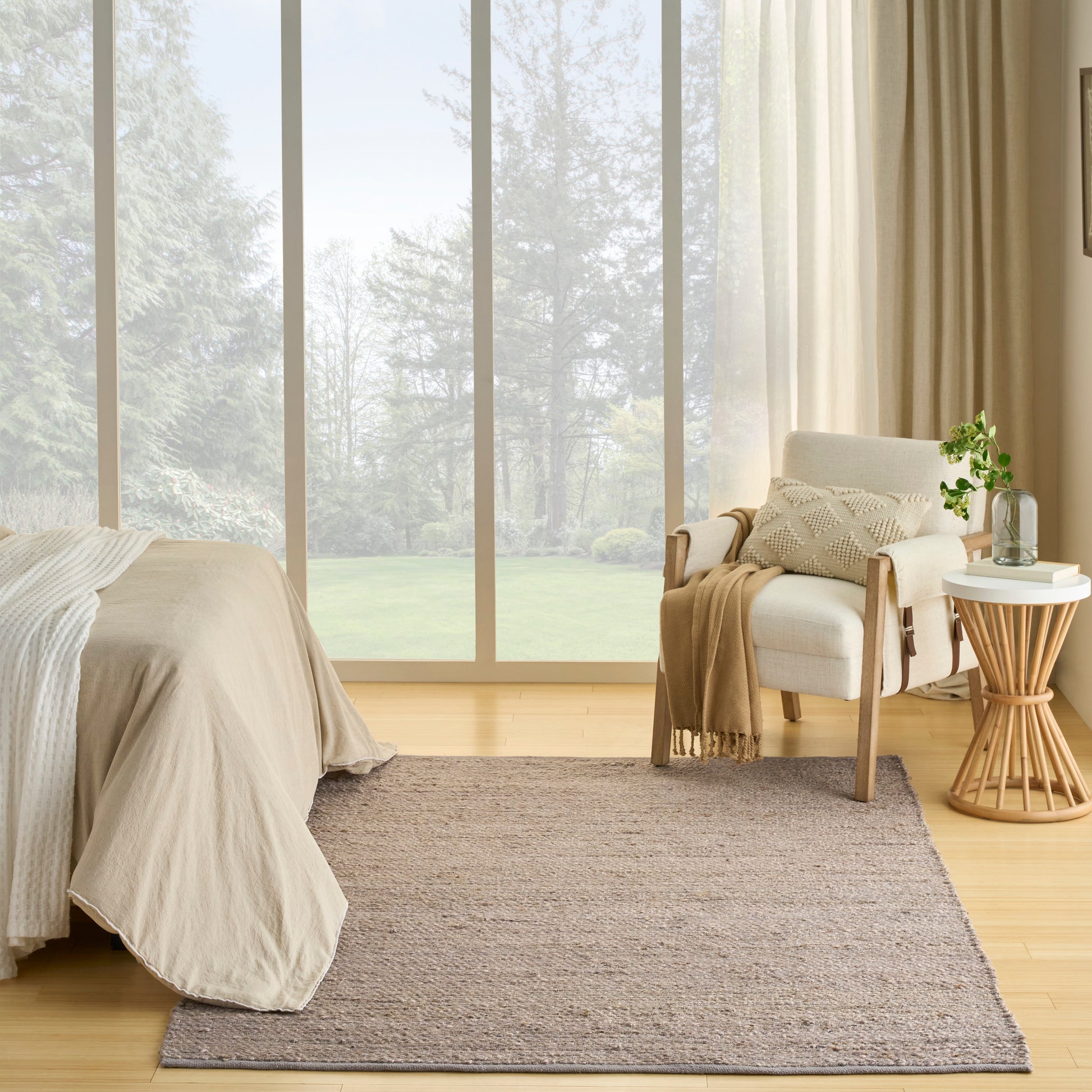 Neutral bedroom with beige bedding, light wood floor, woven rug, cream armchair, and round wood side table by large windows.