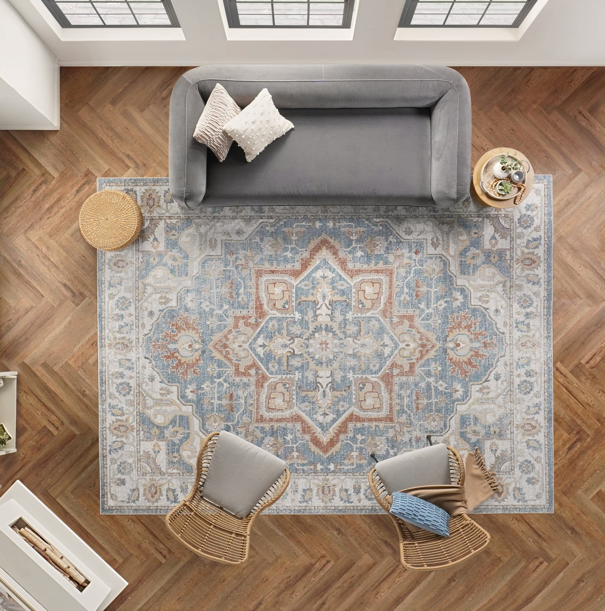 Overhead view: blue/peach patterned rug on herringbone floor, gray sofa, two wicker chairs, round ottoman, plant table.
