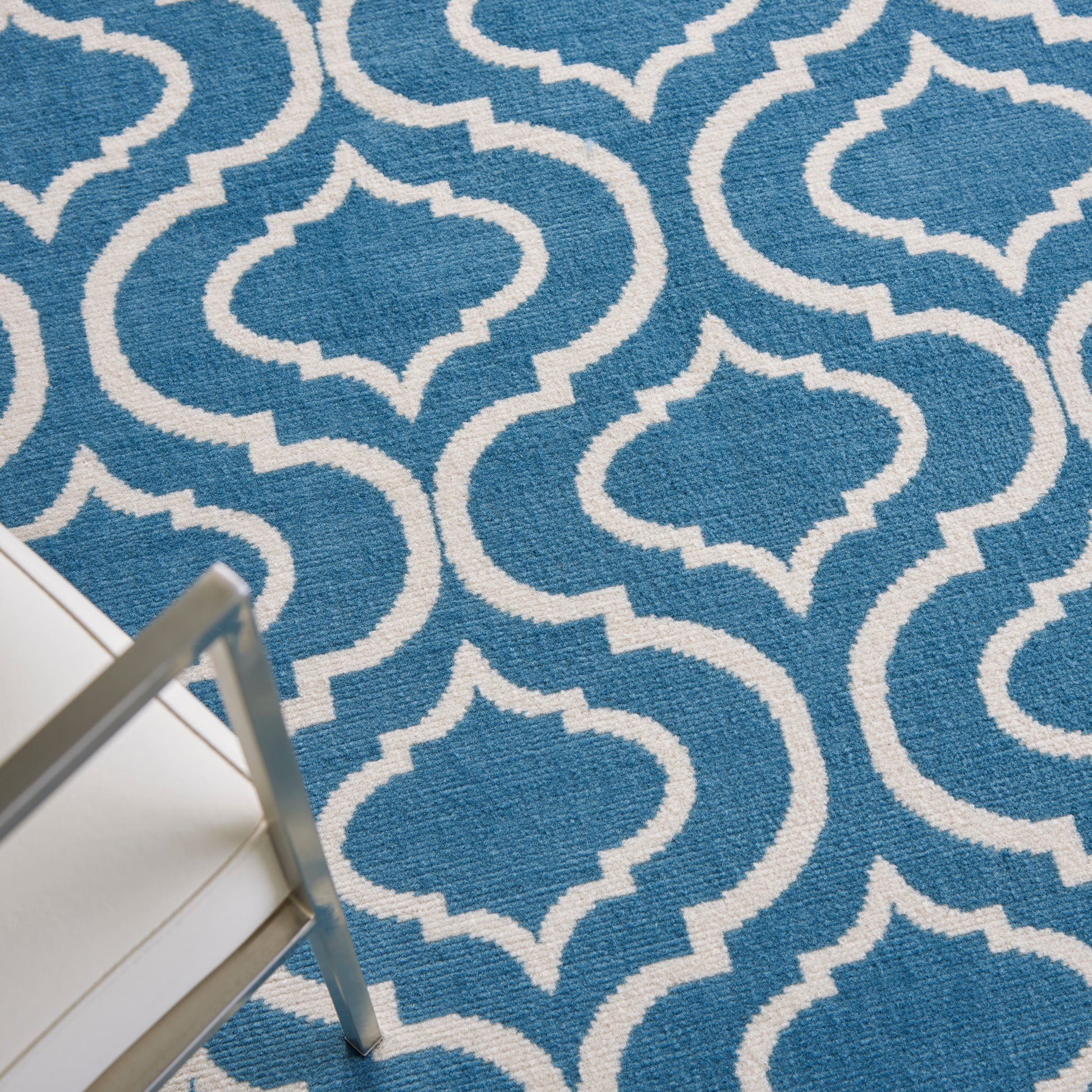 Blue rug with white trellis pattern; textured weave in modern geometric style, with part of white metal chair leg visible.
