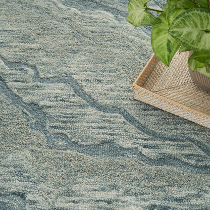 Blue-gray textured rug with an abstract wavy pattern; a natural woven tray holding a green leafy plant.