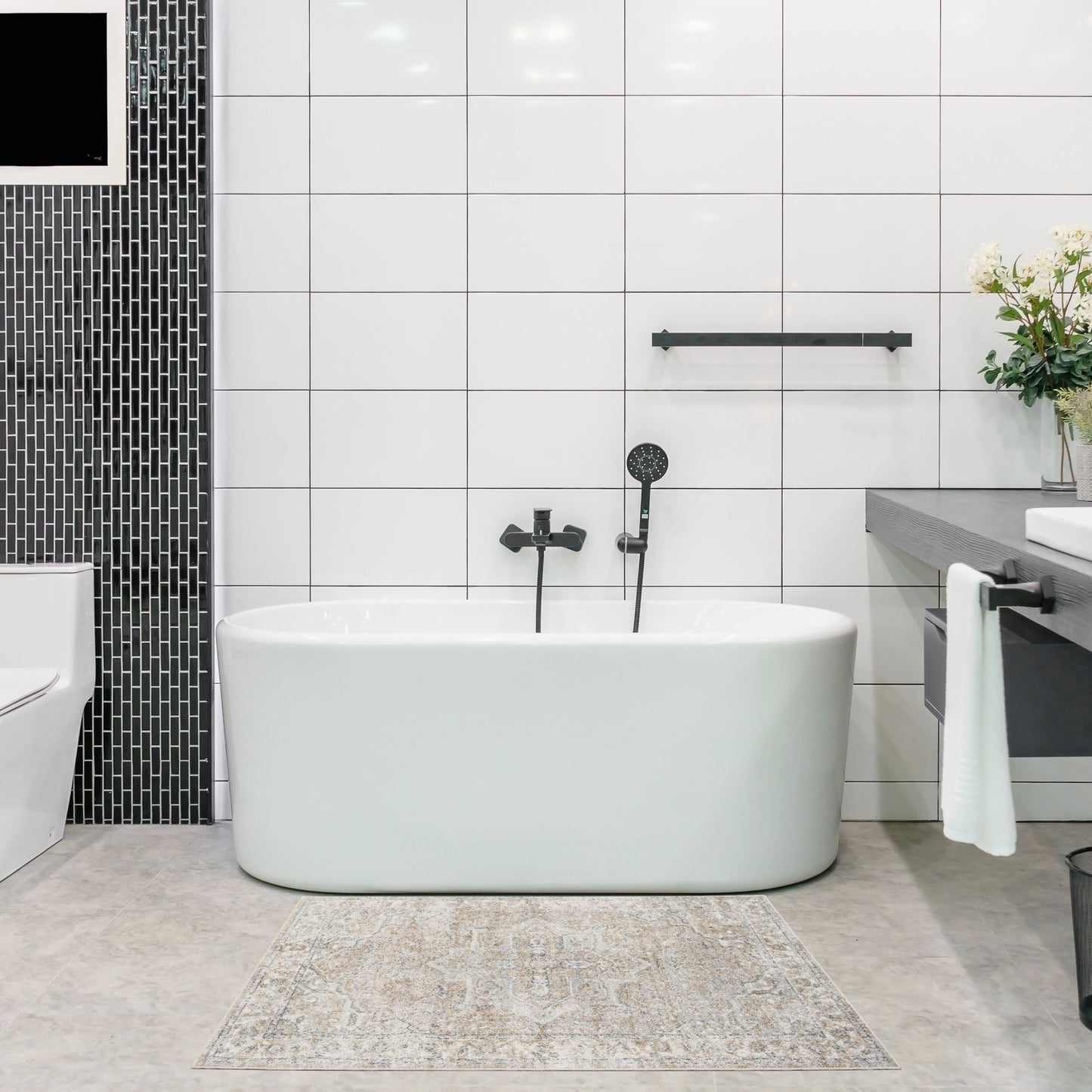 White freestanding oval bathtub with matte black faucet and handheld shower, white tiled walls, black mosaic column, beige patterned rug.