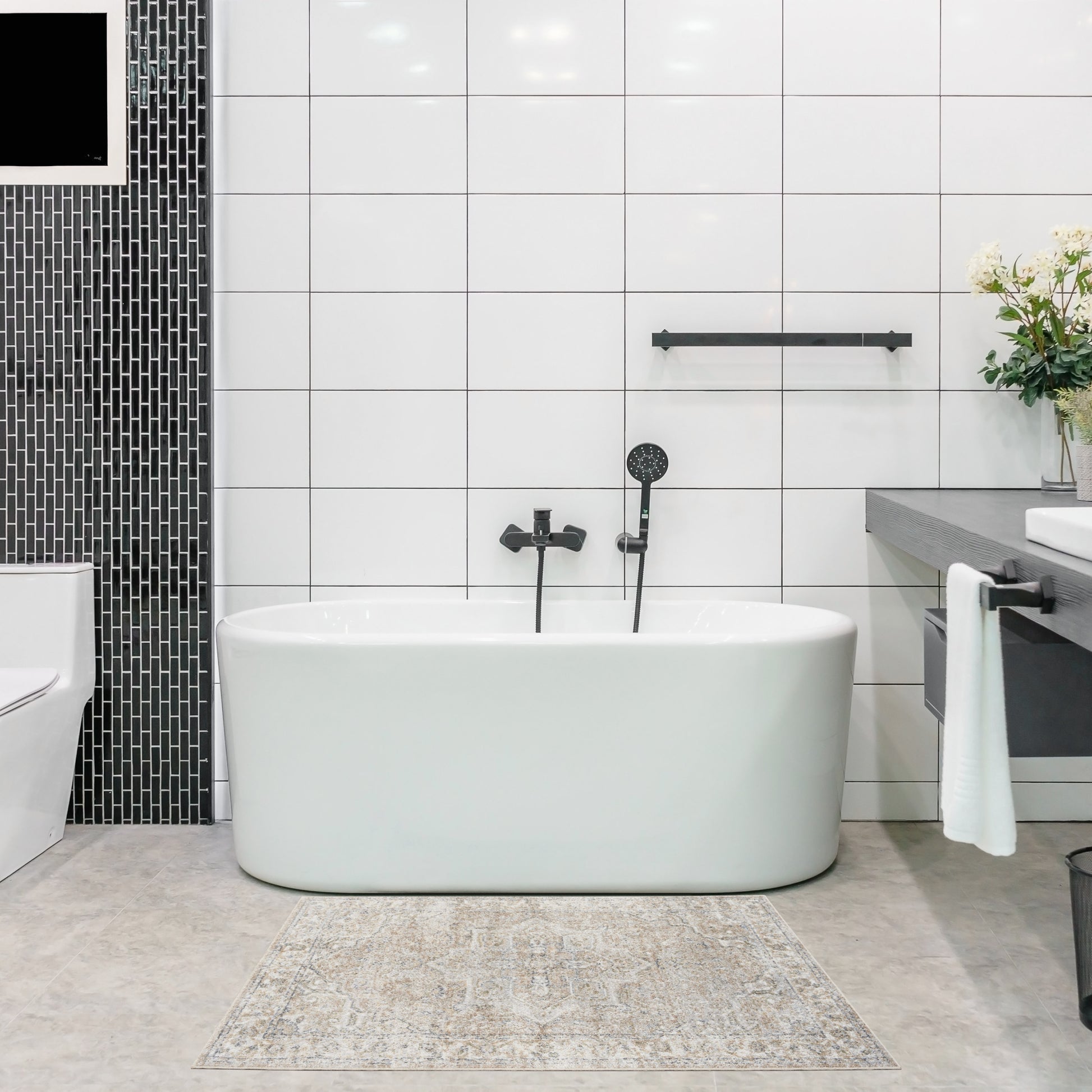White freestanding oval bathtub with matte black faucet and handheld shower, white tiled walls, black mosaic column, beige patterned rug.