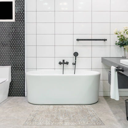 White freestanding oval bathtub with matte black faucet and handheld shower, white tiled walls, black mosaic column, beige patterned rug.
