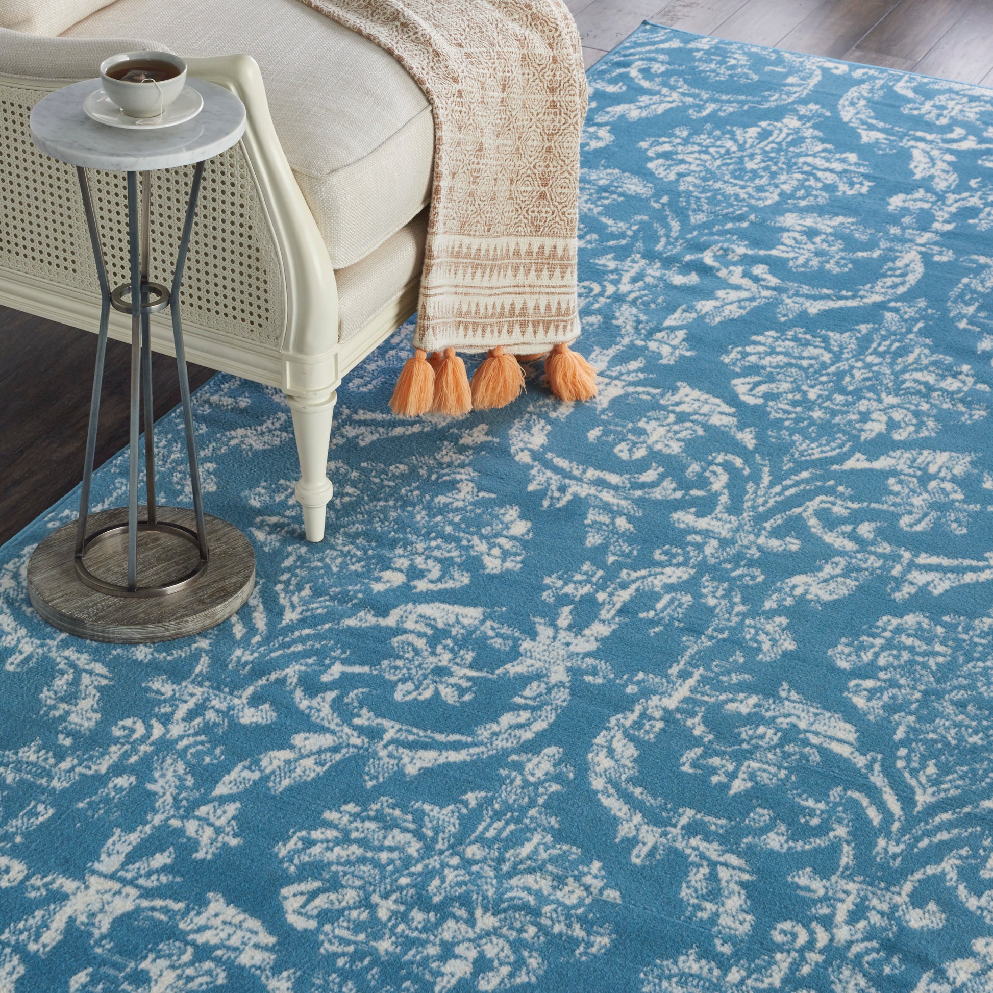 Blue area rug with white ornate scroll pattern, beige sofa, and tassel-trim throw.