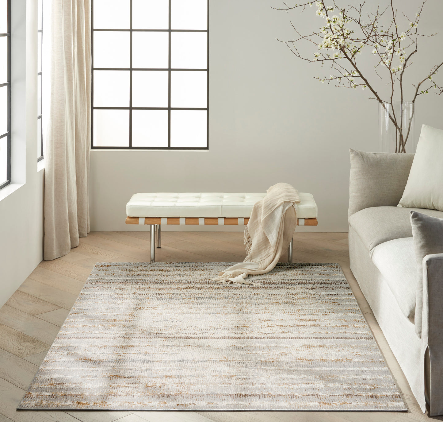 Neutral living room with light beige patterned rug, white tufted bench, beige throw, gray sofa, and branch vase.