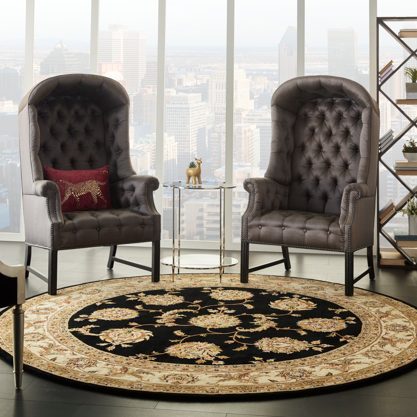 Two tufted gray leather high-back chairs with nailhead trim flank a round black rug with beige floral border and glass side table.