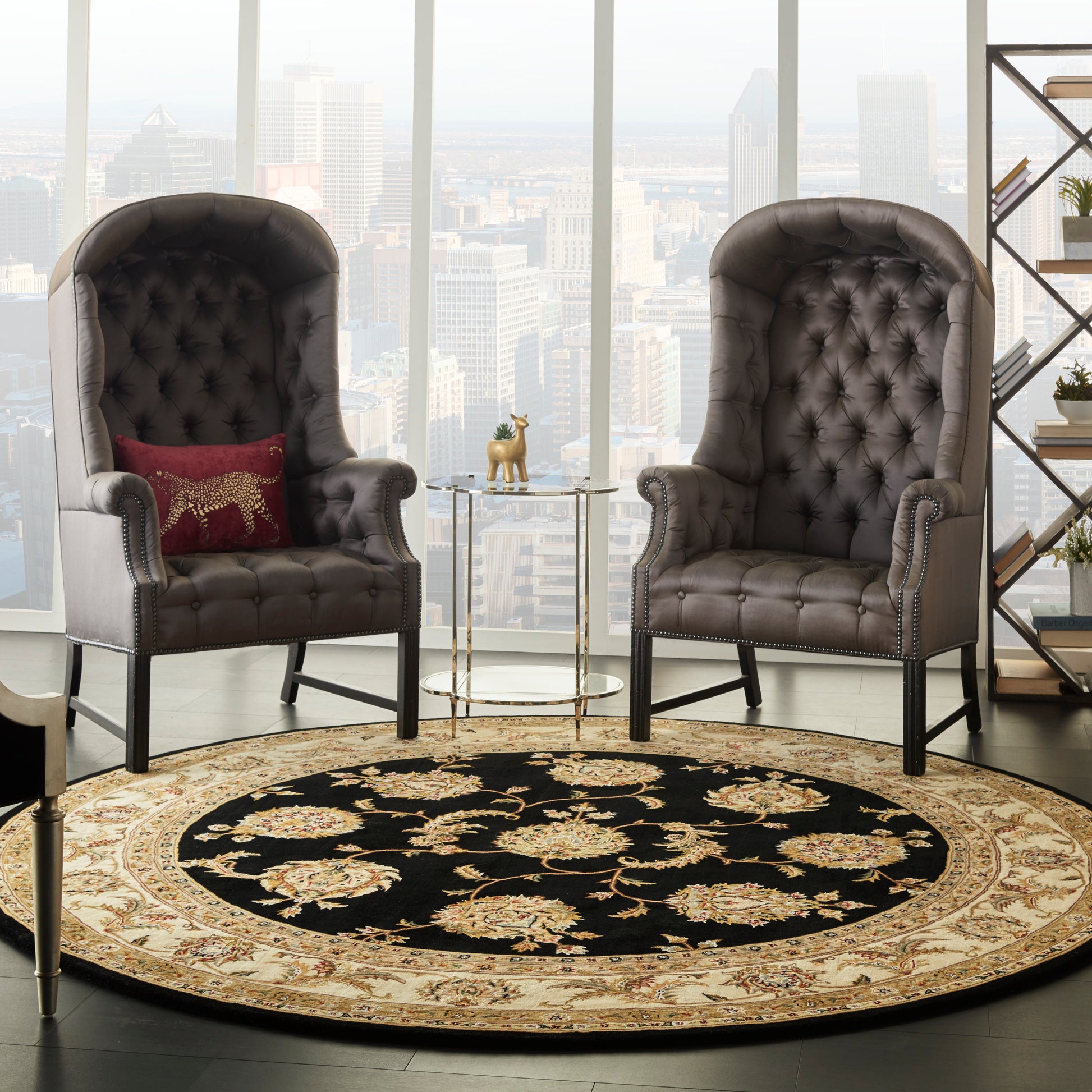 Two tufted gray leather high-back chairs with nailhead trim flank a round black rug with beige floral border and glass side table.
