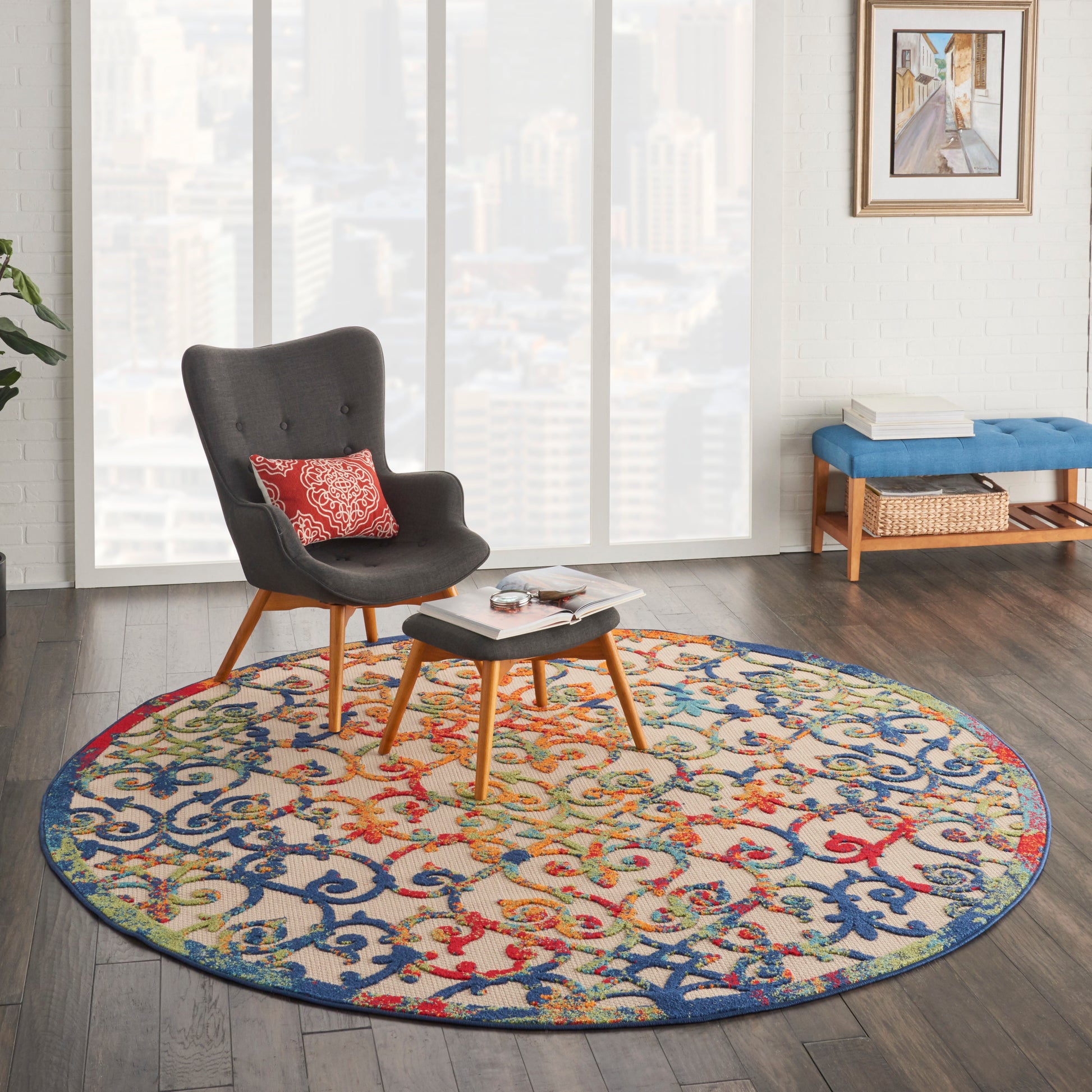 Round multicolor patterned rug under a gray mid-century armchair with wooden legs and ottoman in a bright city living room.