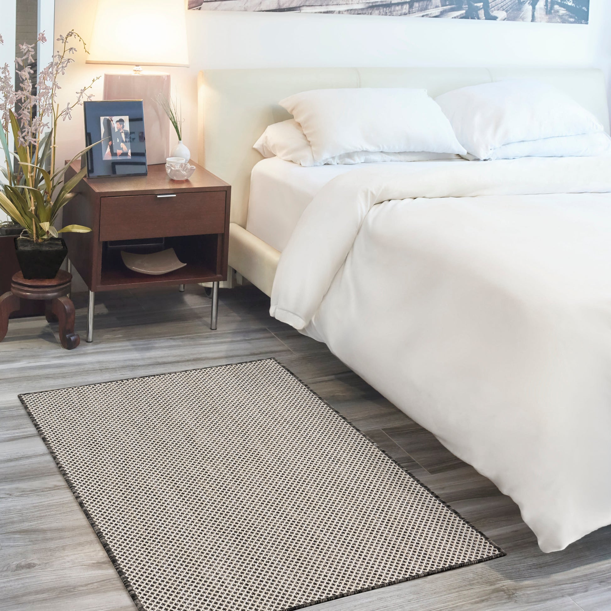 White bedding on a low bed with dark wood nightstand, lamp, framed photo, plant; beige woven rug on grey wood floor.